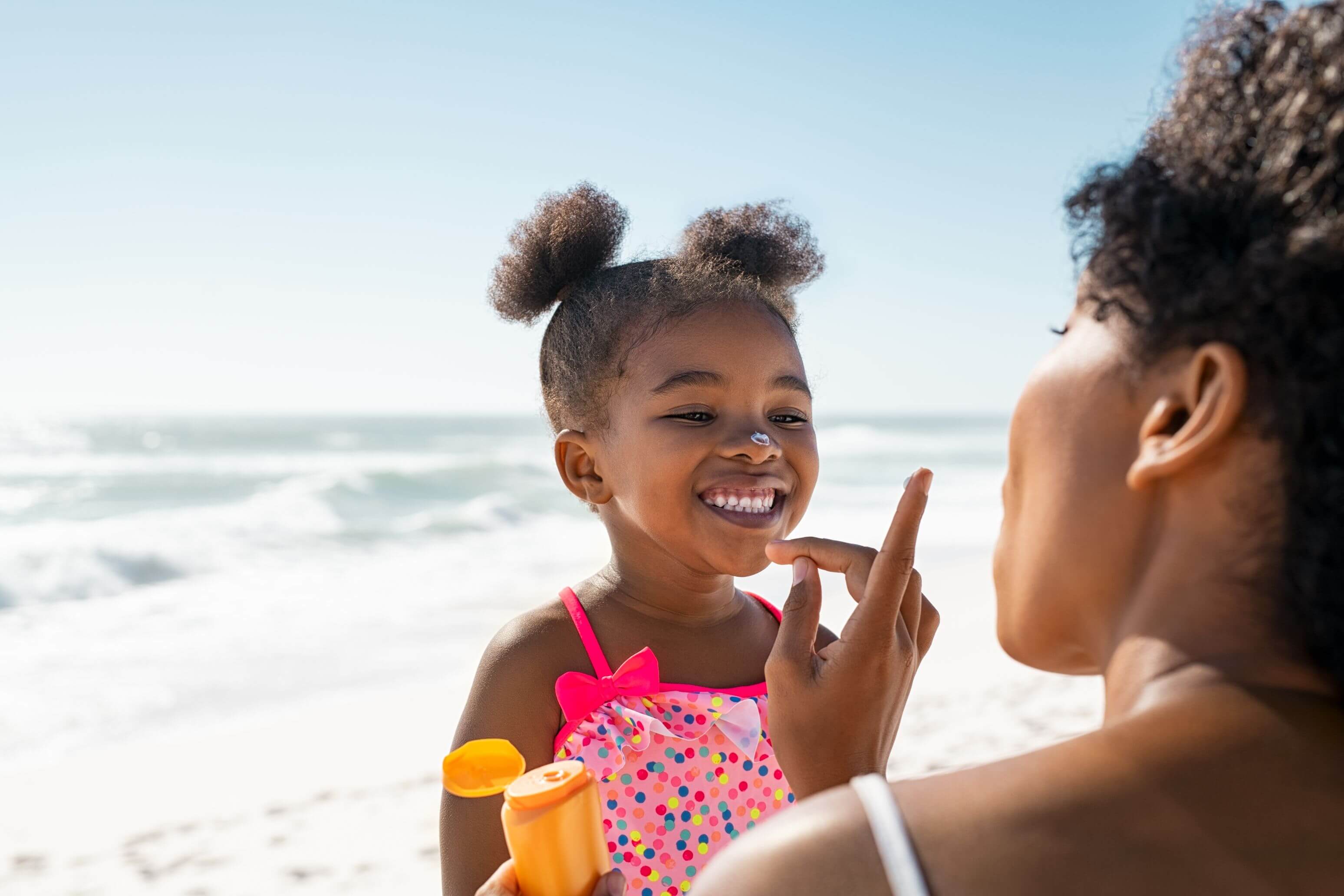 A Black mother applying sunscreen to the face of her daughter at the beach