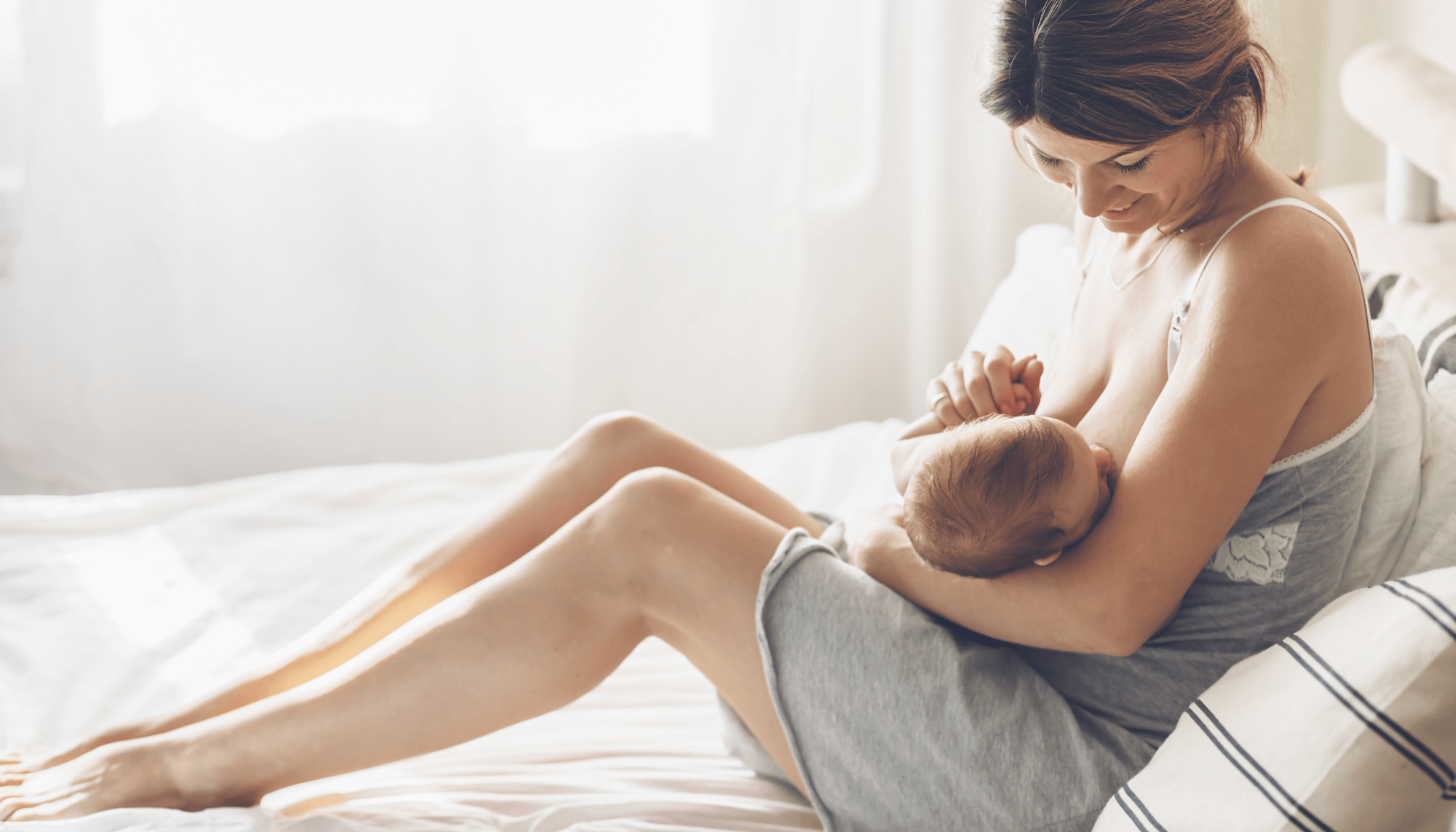 A brown-haired white woman sitting on a bed and breastfeeding her baby