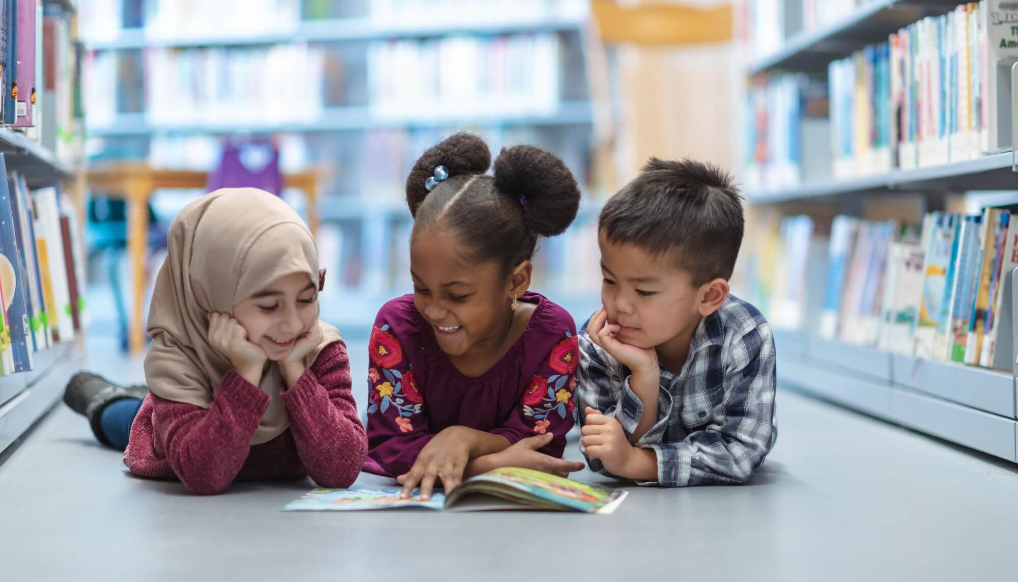 Three five-to-six-year-old children - a Muslim girl, a Black girl, and an Asian boy - lying on the ground of a library and reading a book together.