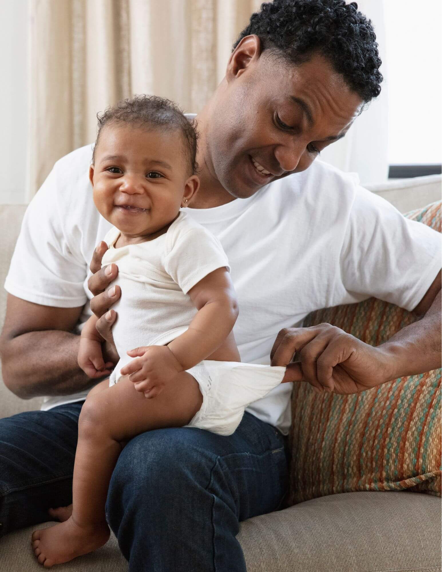 A Black father checking his baby's diaper for poop while the baby sits on his knee. Both are smiling.