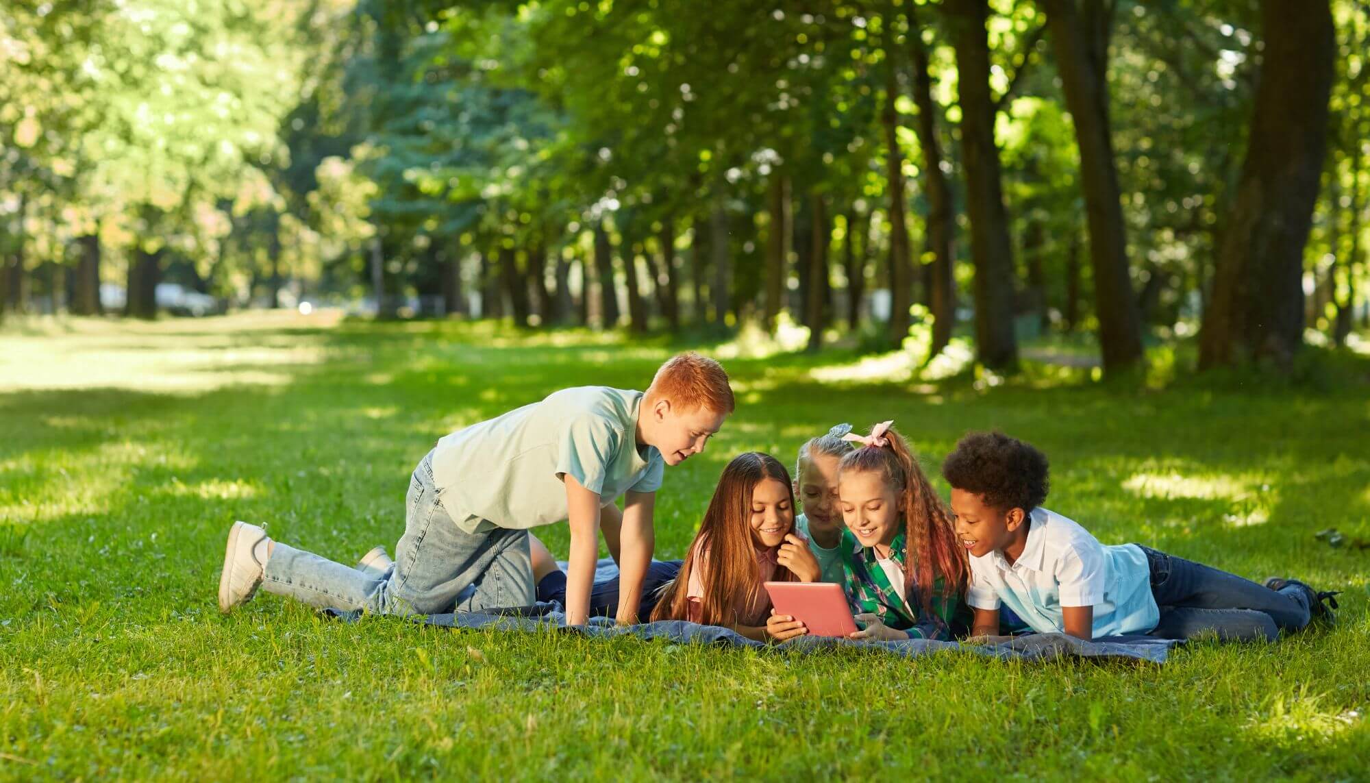 A racially diverse group of teen and tween girls and boys lying on a picnic blanket looking at an iPad. 