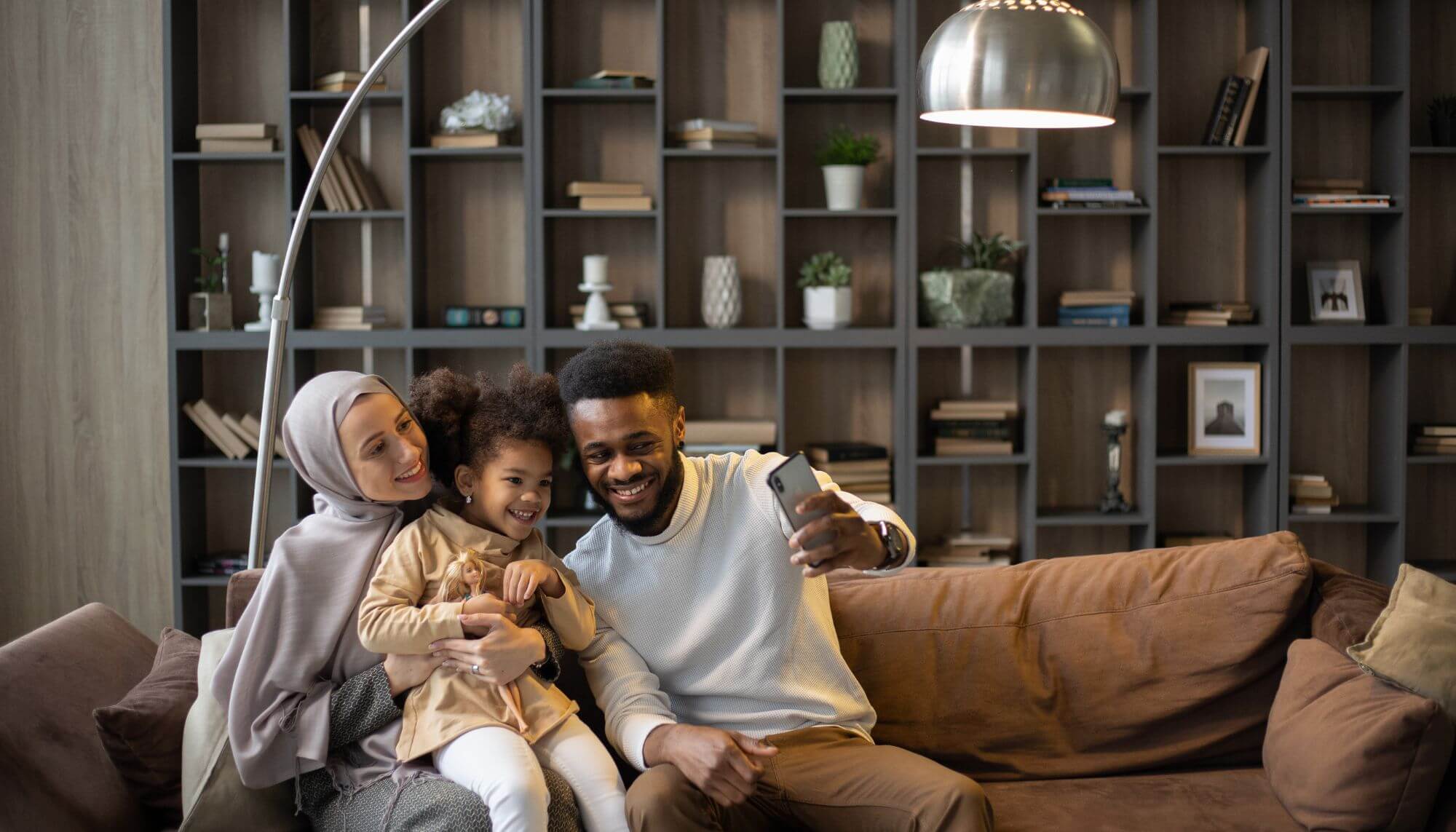 A Muslim mother and Black father sitting together on a sofa with their mixed-race daughter. They are all looking at a phone camera and smiling. 