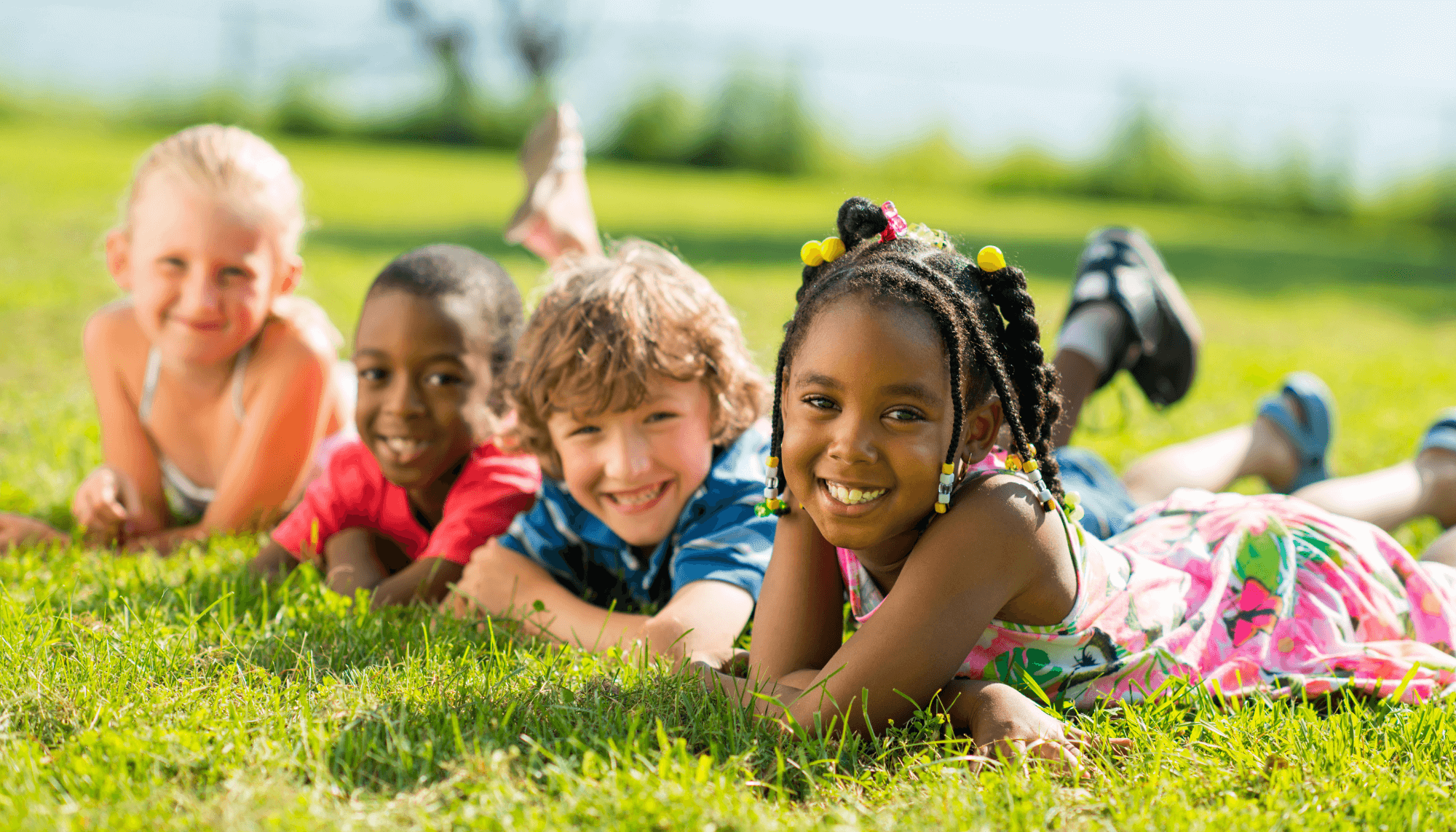 Four six-to-nine-year olds lying on the grass in a line and smiling at the camera: A blonde white girl, a Black boy with shaved hair, a dark blonde boy, and a Black girl with braids. 
