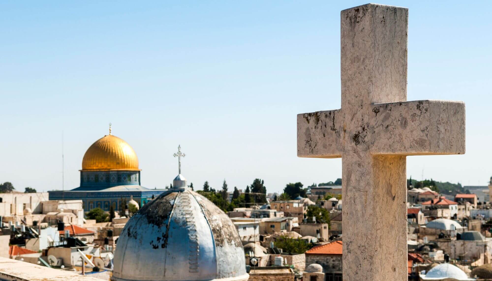 A photo taken of city rooftops showing the cross atop a church and the Qubba of a mosque. 
