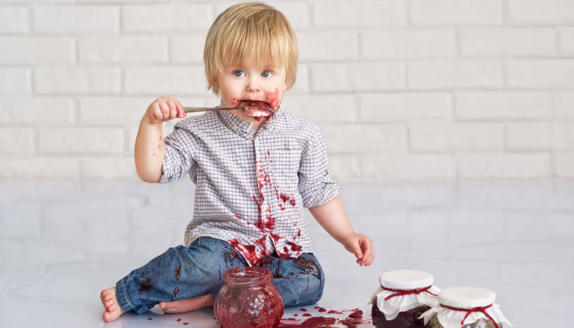A white male toddler eating jam directly from the jar with a large spoon. His face, hands and clothes are covered in jam.
