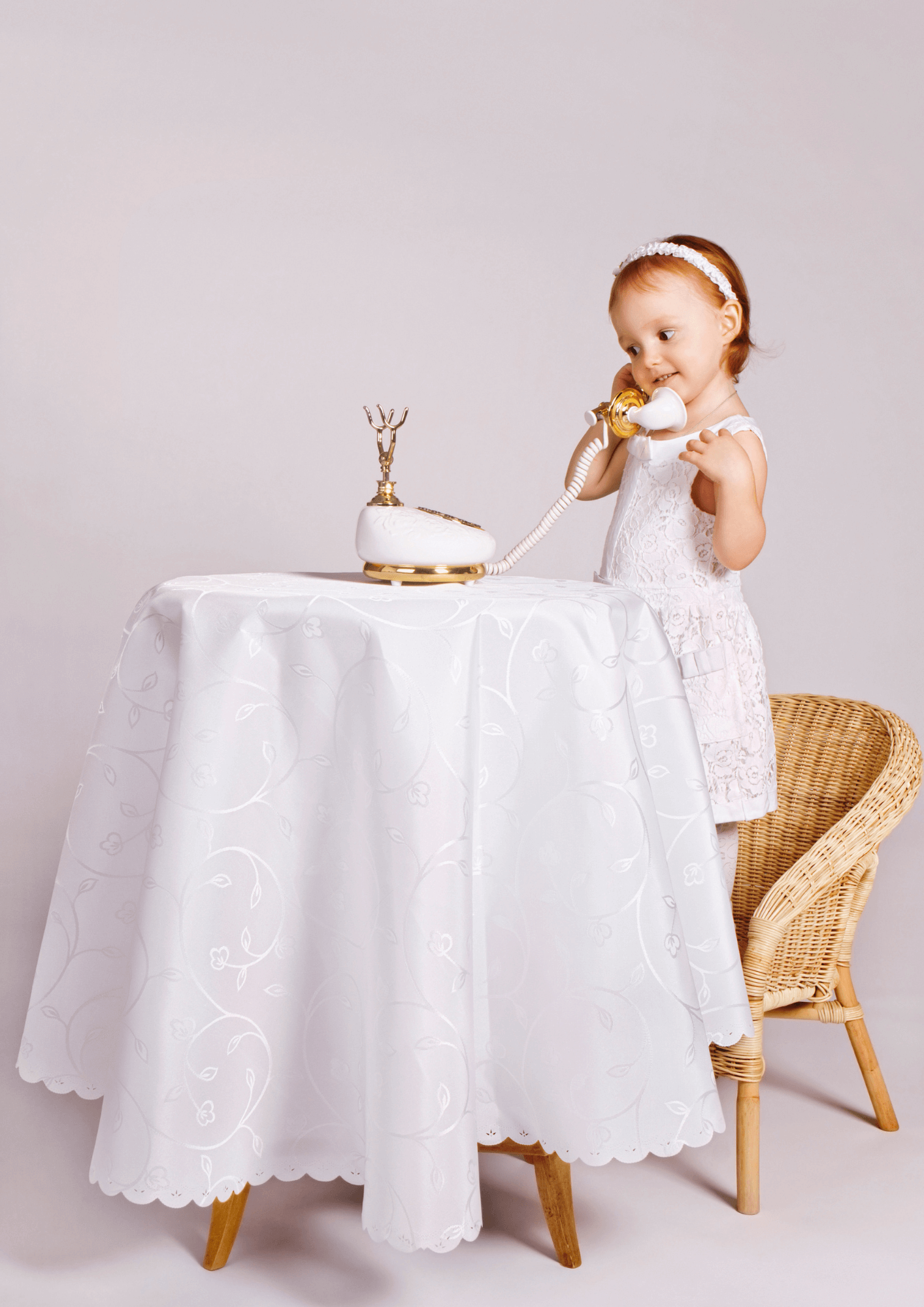 A redheaded toddler standing on a chair to reach a table and pretend to talk on an ornate white and gold telephone