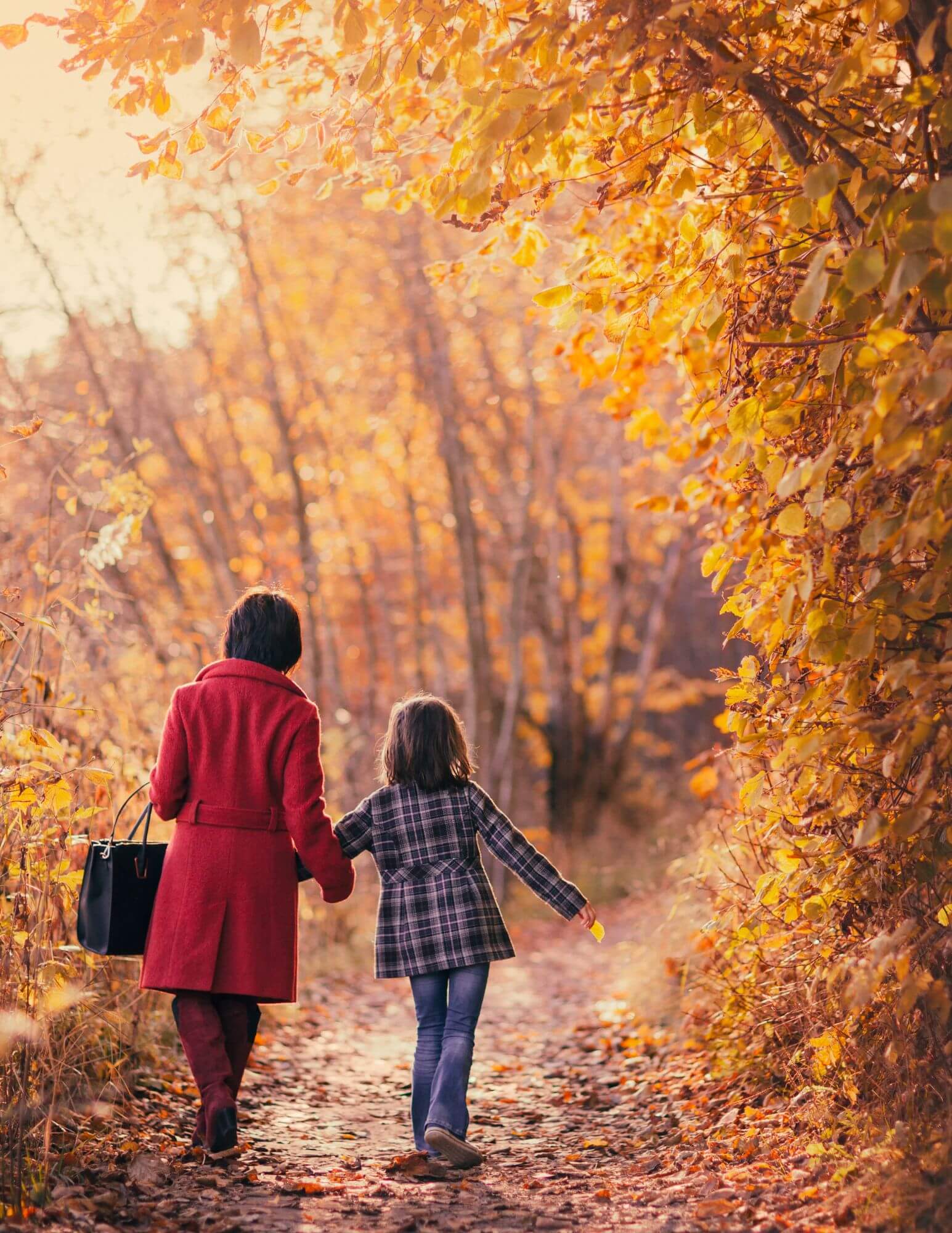 A mother and daughter walking hand in hand through fall leaves away from the camera