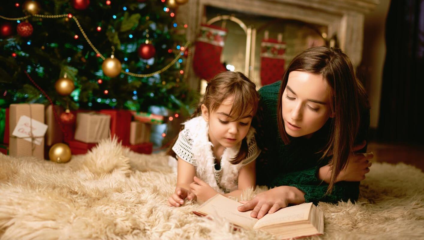 A mother and daughter lying on the floor in front of a Christmas tree and reading a book together.