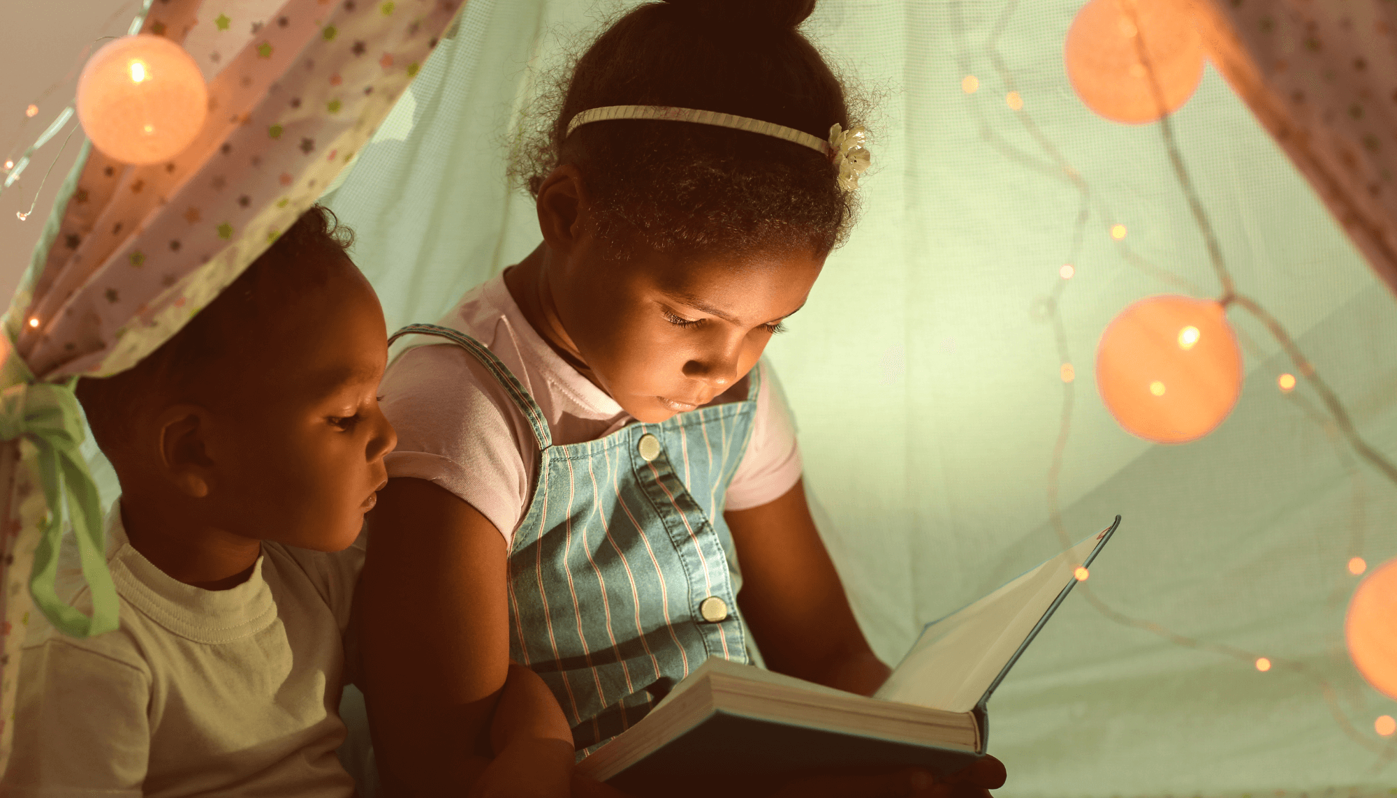 Two children sitting inside a play tent filled with fairy lights and reading a book.