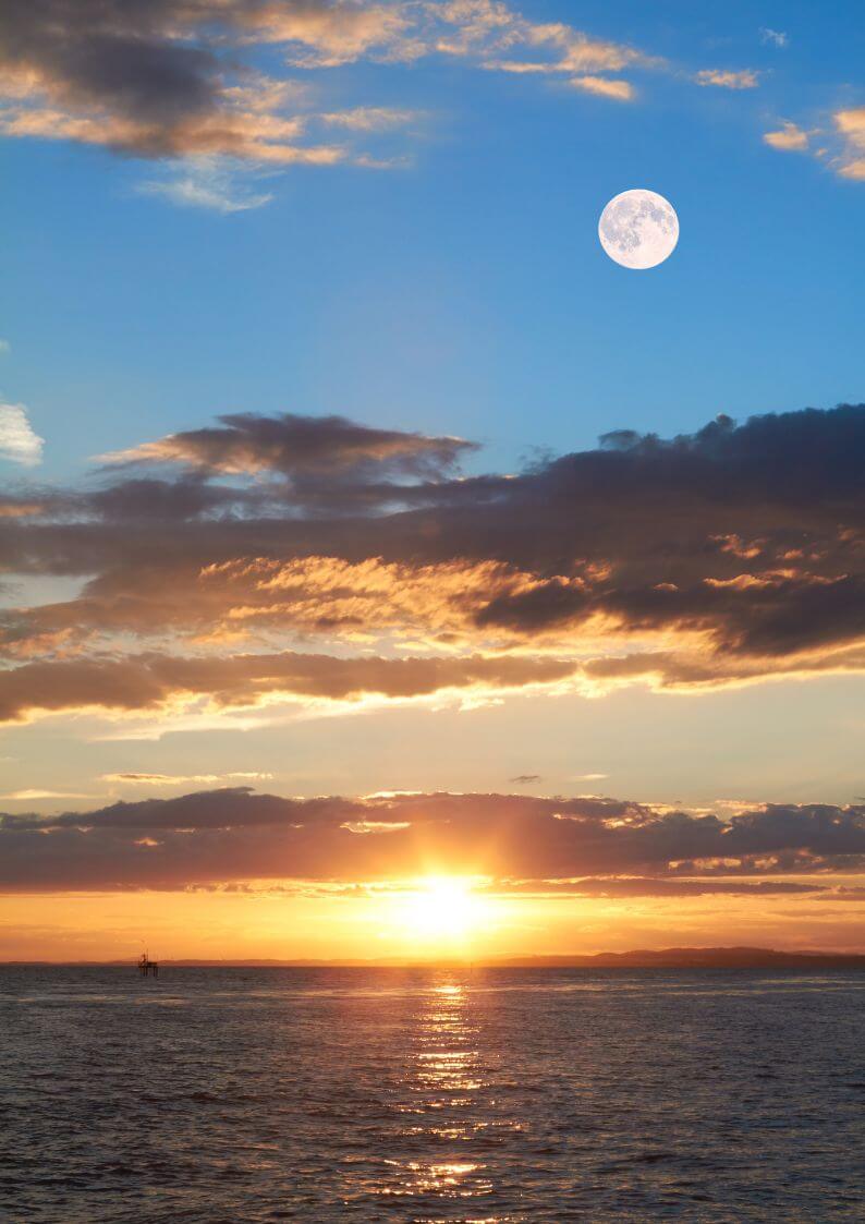 The ocean at sunset, with the moon high in the sky as it turns from blue to orange as the sun sets
