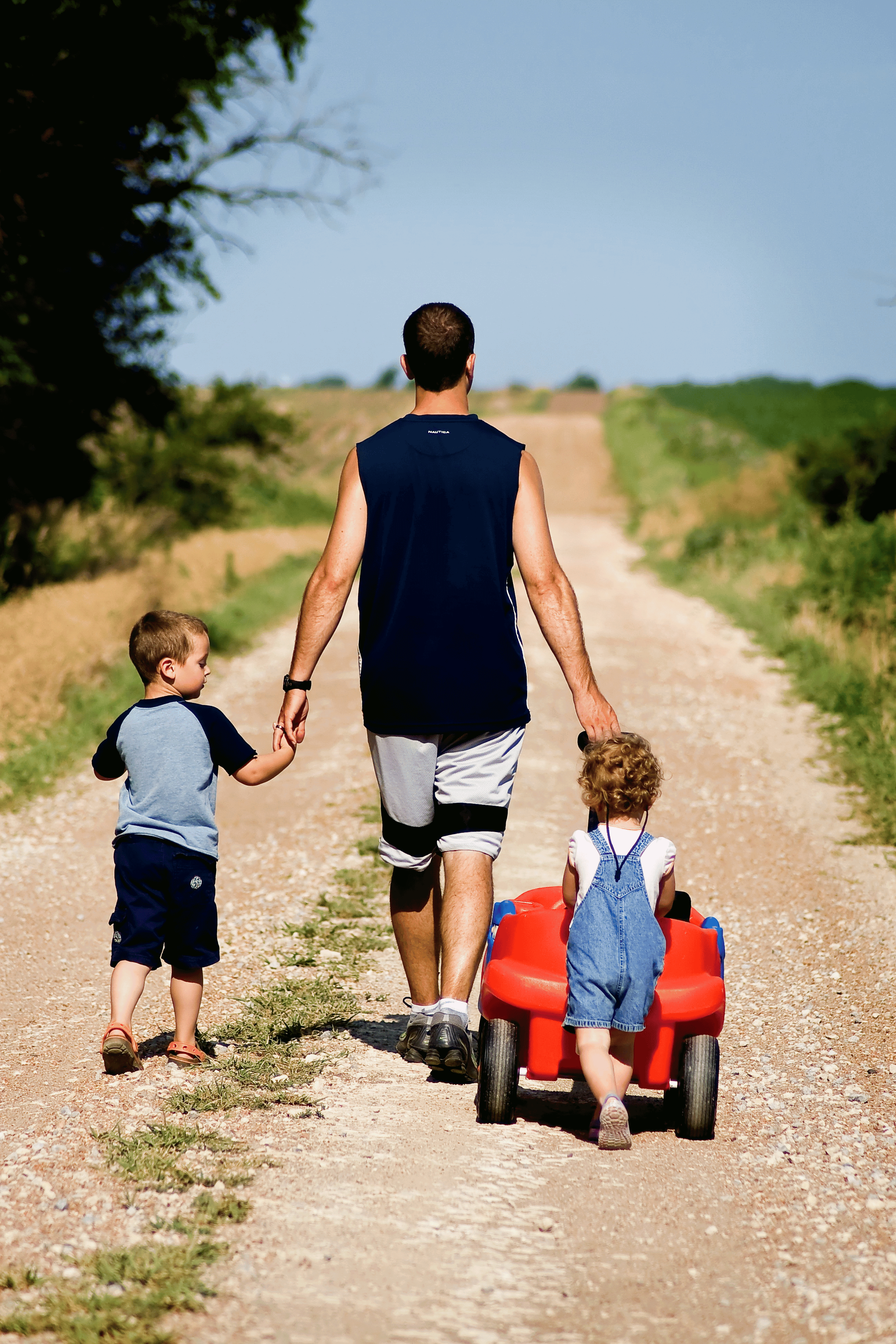 A dad and two children walk away from us along a dirt road