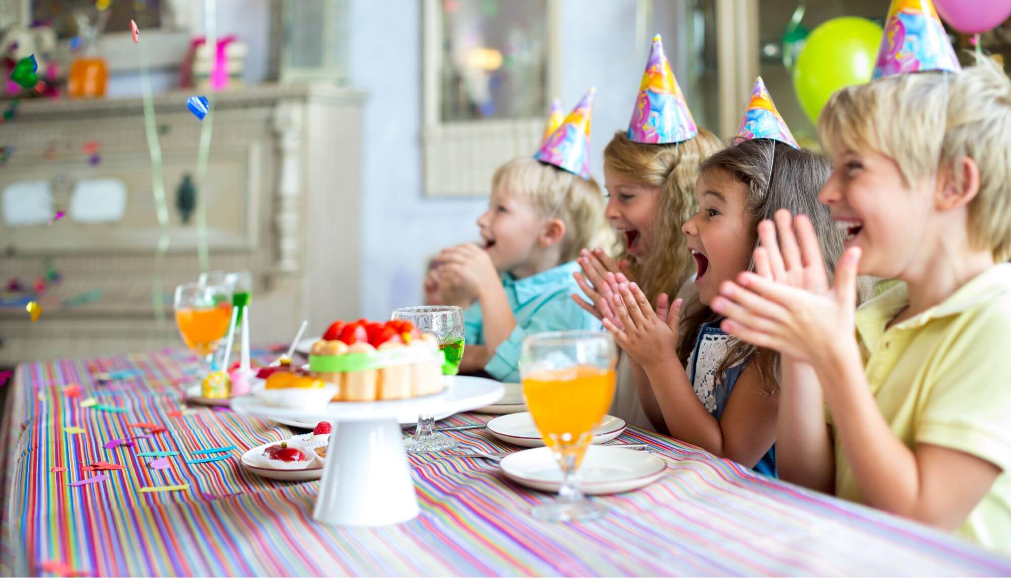 A group of children sitting at a a party table. They are wearing party hats and smiling and clapping.