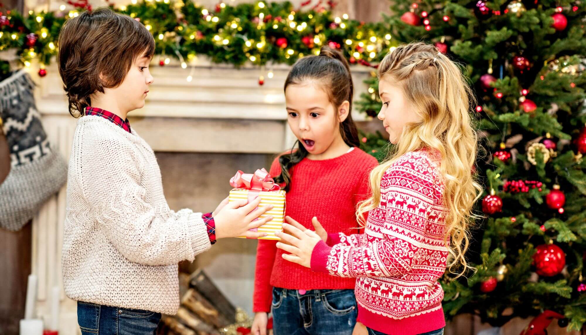 A child handing a wrapped gift to another child as another child looks on in surprise