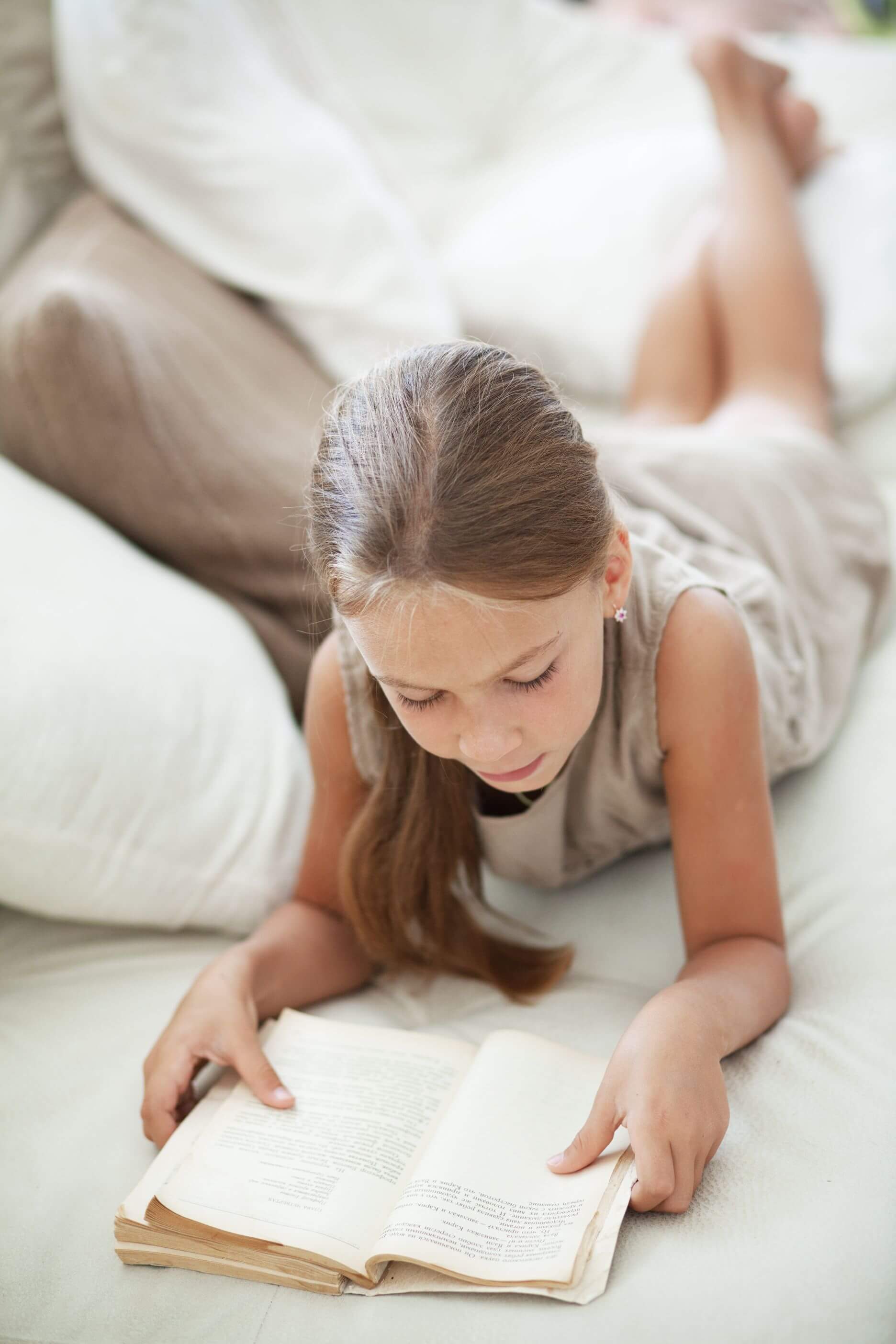 A blonde-haired White girl of around 8 years old lying on her front on a bed reading a book