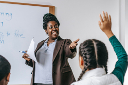 A teacher stands at the front of a class whilst a student raises her hand.