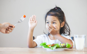 A four-year old Asian girl refusing the cherry tomato being offered to her on a fork by an off-camera adult. She is holding one hand over her mouth and the other in front of the tomato to say "No".