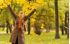 A white woman dressed in a light coat, pants and boots standing under fall trees. She is throwing yellow fallen leaves into the air and she is smiling.