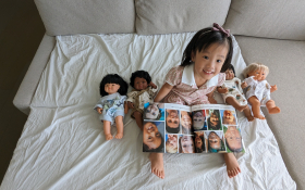 A two-year old Chinese Australian reading a book with photos of many different children in it. She is sitting on a sofa next to four dolls. Each doll has a different skin color.