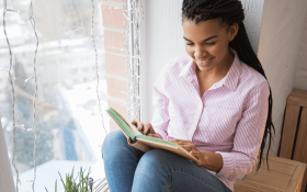 A black woman sitting in a window reading nook and reading a book