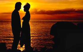 The silhouttes of a man and woman standing close to each other in front on an orange sunset over water