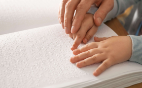An adult's hand guiding a pair of child's hands across the pages of a book printed in Braile.