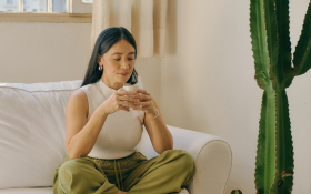 A woman sits cross legged on a couch by a cactus.